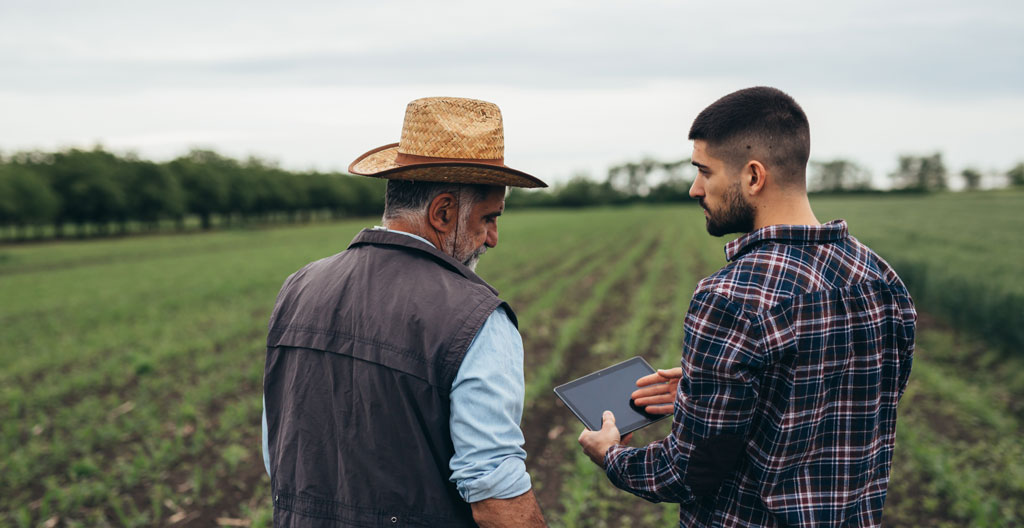 Deux agriculteurs discutant d'un outil de gestion numérique devant un bâtiment agricole, l'un montrant l'écran de son smartphone